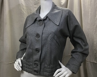 May include: A gray wool jacket with a wide collar and button closure. The jacket features long sleeves, a tailored fit, and decorative stitching along the bottom. The jacket is displayed on a mannequin.