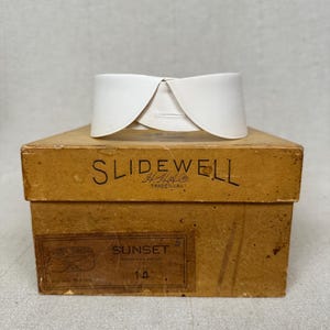 Könnte beinhalten: Ein weißer, abnehmbarer Hemdkragen auf einem beigen Pappkarton. Die Schachtel trägt die Aufschrift SLIDEWELL sowie ein Etikett mit dem Wort SUNSET und der Zahl 14. Der Kragen hat ein geschwungenes, überlappendes Design mit einem Knopfloch.