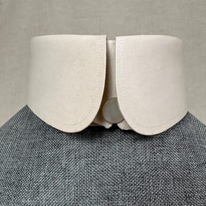 Puede incluir: Un cuello desmontable de color crema con forma redondeada. El cuello está hecho de una tela resistente y se sujeta con un botón. El cuello está colocado sobre una chaqueta de tweed gris.