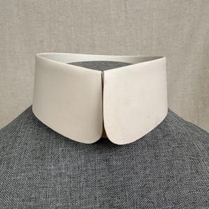 Op de afbeelding: Een stijve, gebroken witte afneembare kraag met een afgerond, hoogstaand ontwerp. De kraag heeft een gladde, matte afwerking en een strakke, gebogen rand die in het midden aan de voorzijde samenkomt voor een gestructureerde, formele uitstraling.