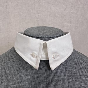 May include: A detachable white shirt collar made of a textured fabric. The collar features two pointed tips, each secured with a small white button. The collar is displayed on a grey fabric-covered mannequin form, highlighting the crisp white finish.