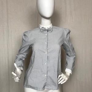 Puede incluir: Una camisa de manga larga a rayas blancas y azules con una pajarita blanca. La camisa tiene un cierre de botones en la parte delantera y una silueta ajustada.