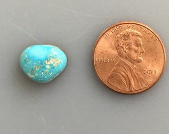May include: A turquoise cabochon gemstone in a heart shape, next to a US penny for size comparison. The turquoise is a light blue color with brown and white matrix.
