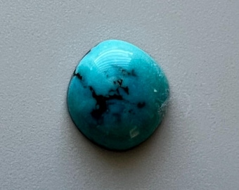 May include: A polished, oval-shaped turquoise gemstone with a vibrant blue-green hue. The stone features dark, irregular veining patterns. The gemstone is set against a neutral gray background.