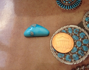 May include: A polished, triangular turquoise stone with gray inclusions. A copper one-cent coin is next to the stone for size comparison. The stone is set against a brown background with Native American jewelry.