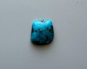 May include: A polished turquoise gemstone with a rounded rectangular shape. The stone features a vibrant blue hue with dark matrix patterns and a glossy surface. The gemstone is set against a neutral background.