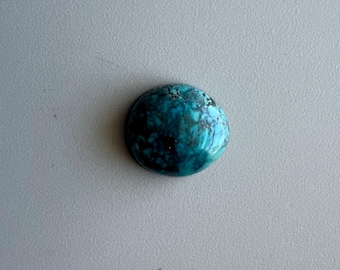 May include: A round, polished turquoise gemstone with a mottled pattern of deep teal, dark blue, and light blue hues. The stone has a glossy, reflective surface, and is set against a neutral gray background.