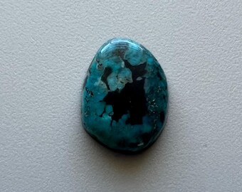 May include: A polished, oval-shaped gemstone with a striking mix of teal and black colors. The stone's surface is smooth and glossy, with a pattern of swirling teal and dark black patches. The gemstone is set against a light gray background.