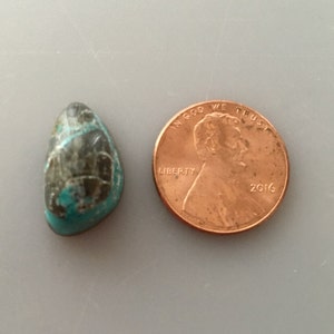 May include: A polished, teardrop-shaped gemstone with a mix of teal and dark gray colors, next to a copper penny. The penny is dated 2016 and is used for size comparison. The gemstone has a smooth, glossy surface.