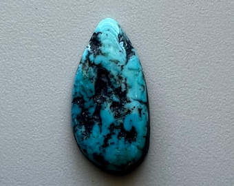 May include: A teardrop-shaped turquoise gemstone with a polished surface. The stone features a vibrant blue hue with dark, irregular black veining throughout. The gemstone is set against a neutral gray background.