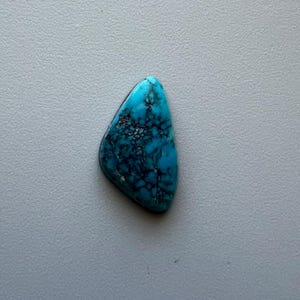Puede incluir: Una piedra preciosa de turquesa pulida en forma de lágrima, con un vibrante tono azul y un intrincado veteado negro. La piedra se encuentra sobre un fondo gris neutro, resaltando su belleza natural y su patrón único.