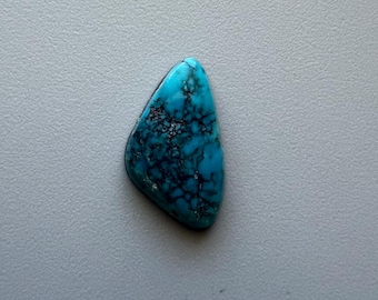 May include: A polished, teardrop-shaped turquoise gemstone with a vibrant blue hue and intricate black veining. The stone is set against a neutral gray background, highlighting its natural beauty and unique pattern.