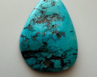 May include: A polished, teardrop-shaped turquoise gemstone with a vibrant blue-green hue. The stone features intricate dark brown matrix patterns throughout its surface, creating a unique and natural design. The gemstone is smooth and glossy.