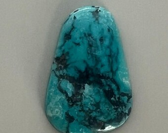 May include: A polished, teardrop-shaped turquoise gemstone with dark, irregular veining. The stone's surface is a vibrant teal color, with areas of lighter blue and black inclusions. The gemstone is set against a neutral gray background.
