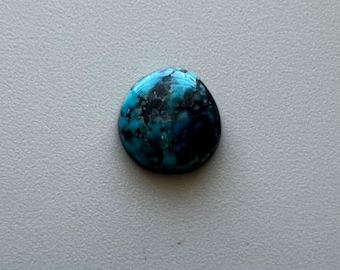 May include: A polished, oval-shaped gemstone with a striking blend of turquoise and deep blue hues, accented by dark, mottled patterns. The stone is set against a neutral gray background, highlighting its natural beauty.