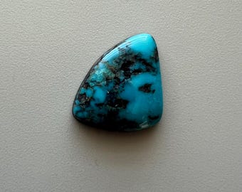 May include: A polished turquoise gemstone with a triangular shape. The stone features a vibrant blue color with dark, black matrix patterns. The surface is smooth and glossy, reflecting light. This gemstone is suitable for jewelry making.