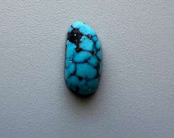 May include: A polished turquoise gemstone with a striking matrix of black veins. The stone is oval-shaped and displays a vibrant blue-green color. The gemstone is set against a neutral gray background.