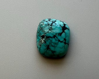May include: A polished, turquoise gemstone with a rounded rectangular shape. The stone features a striking matrix of black veining across its surface, creating a unique pattern. The gemstone is set against a neutral background.
