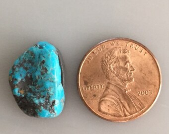 May include: A turquoise stone with a dark blue and green color, next to a US penny for size comparison. "In God We Trust" and "Liberty" are visible on the coin.