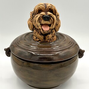 May include: A brown ceramic dog-themed container with a lid. The dog's head is sculpted on top, with a happy expression and its tongue sticking out. The container has a glossy finish and two small handles on the sides.