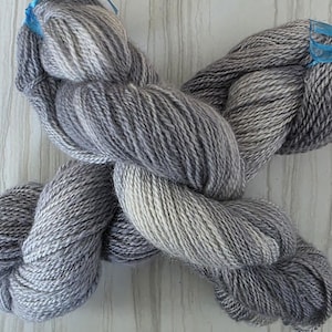 May include: A skein of gray yarn with varying shades of light and dark gray. The yarn is twisted and tied with a blue ribbon. The yarn appears soft and is likely made of wool or a similar fiber. Ideal for knitting or crocheting projects.