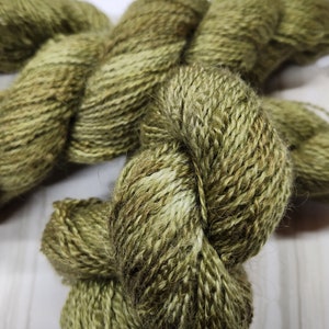 May include: Close-up of a skein of olive green yarn. The yarn has a textured appearance with variations in color, including lighter and darker shades of green and brown. The yarn is tightly wound, suggesting it is ready for knitting or crocheting projects.