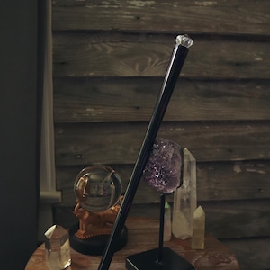 Peut inclure: Une baguette noire avec un dessus en cristal est posée contre une table en bois. Une boule de cristal transparente, une améthyste violette et des cristaux de quartz sont disposés sur la table, suggérant un thème mystique ou spirituel.