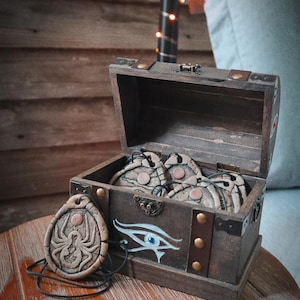 Può includere: Un piccolo forziere di legno aperto con un design decorativo dell'Occhio di Horus e un ciondolo abbinato. Il forziere contiene diversi oggetti rotondi color beige con accenti color rame. Il ciondolo presenta un disegno di ragno.