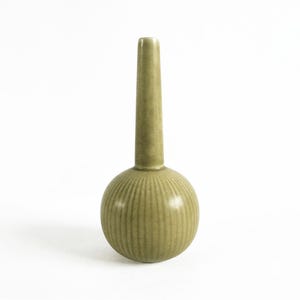 Puede incluir: Un jarrón de cerámica verde salvia con base redonda y cuello cilíndrico alto. La base tiene detalles acanalados verticales. El jarrón es un elemento decorativo, adecuado para exhibir flores o como pieza de arte independiente.