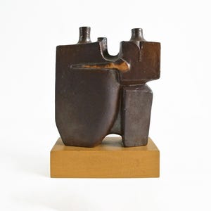 Op de afbeelding: Abstract bronzen sculptuur met een geometrisch ontwerp. De sculptuur heeft twee flesachtige vormen die zijn verbonden door een horizontaal element, gemonteerd op een rechthoekige houten basis. De sculptuur is donkerbruin van kleur.