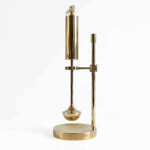 Puede incluir: Una lámpara de aceite vintage de latón con un depósito cilíndrico, un tallo largo y una base redonda. La lámpara tiene un diseño único con un pequeño elemento esférico suspendido debajo del depósito. La lámpara es de latón brillante y se alza sobre un fondo blanco.