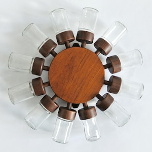 Puede incluir: Un especiero de madera con diseño circular, con un disco central de madera y doce botes de especias de vidrio transparente. Los botes están dispuestos en un patrón radial, cada uno sujeto por un soporte de madera marrón oscuro. Fondo blanco.