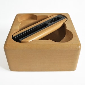 May include: A wooden nutcracker with a black metal cracking mechanism, set in a matching wooden base. The base features two curved indentations. The nutcracker and base are made of light-coloured wood.