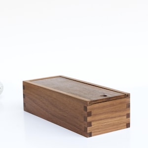 May include: A rectangular wooden box with a lid, made of dark brown wood with a natural wood grain. The box has a small hole in the lid.