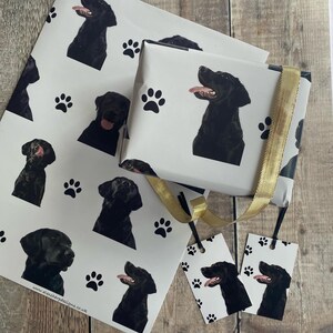 May include: Black Labrador Retriever wrapping paper with paw prints. The paper is white with black paw prints and black Labrador Retriever heads. The paper is wrapped around a gift box with a gold ribbon. There are two gift tags with the same design as the wrapping paper.