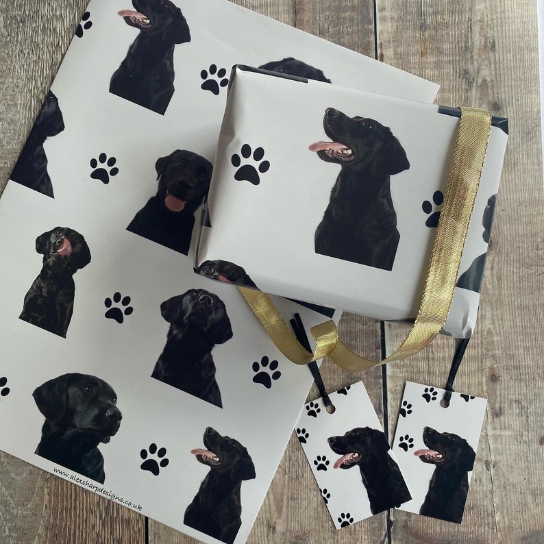 May include: Black Labrador Retriever wrapping paper with paw prints. The paper is white with black paw prints and black Labrador Retriever heads. The paper is wrapped around a gift box with a gold ribbon. There are two gift tags with the same design as the wrapping paper.