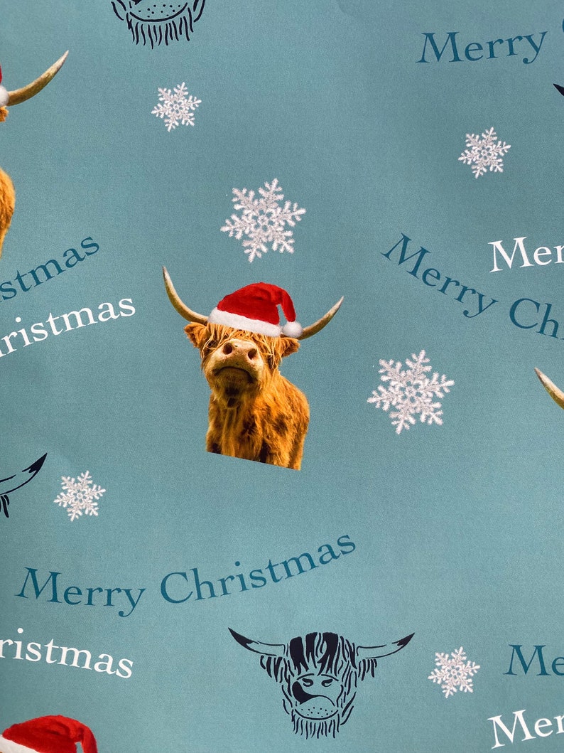 Op de afbeelding: Blauw en wit kerstpapier met een herhalend patroon van Schotse Hooglanders met kerstmanmutsen, sneeuwvlokken en de tekst "Merry Christmas".