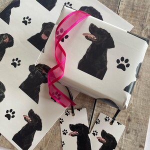 May include: White gift wrap with a black Labrador Retriever design. The wrap features paw prints and a pink ribbon. There are also two gift tags with the same design.