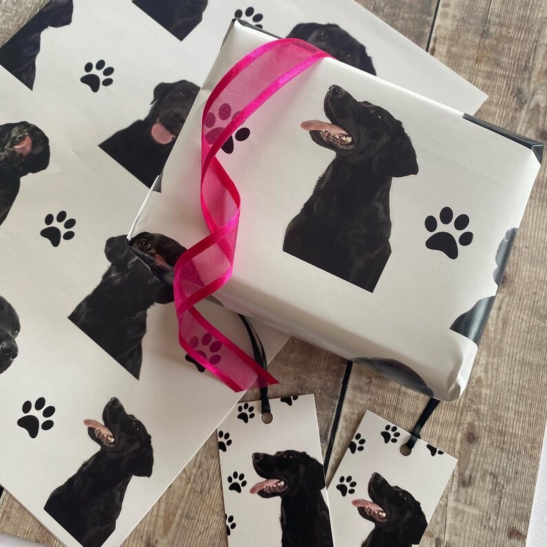 May include: White gift wrap with a black Labrador Retriever design. The wrap features paw prints and a pink ribbon. There are also two gift tags with the same design.