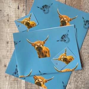 K&ouml;nnte beinhalten: Blaues Geschenkpapier mit einem sich wiederholenden Muster aus schwarzen und wei&szlig;en Highland-Kuh-Silhouetten und vollfarbigen Highland-Kuh-Bildern. Das Papier ist gefaltet, um einen Geschenkanh&auml;nger mit einem vollfarbigen Highland-Kuh-Bild zu zeigen.
