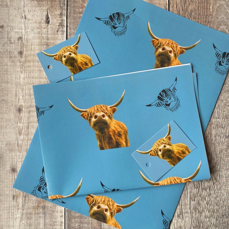 K&ouml;nnte beinhalten: Blaues Geschenkpapier mit einem sich wiederholenden Muster aus schwarzen und wei&szlig;en Highland-Kuh-Silhouetten und vollfarbigen Highland-Kuh-Bildern. Das Papier ist gefaltet, um einen Geschenkanh&auml;nger mit einem vollfarbigen Highland-Kuh-Bild zu zeigen.