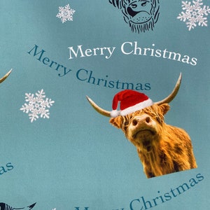Op de afbeelding: Een blauw cadeaupapierontwerp met een Schotse hooglander die een kerstmanmuts draagt en de tekst "Merry Christmas" die zich herhaalt.
