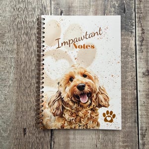 May include: A spiral-bound notebook featuring a Cockapoo dog illustration. The cover reads "Impawtant Notes" with a paw print. The dog is golden-brown. The notebook is on a wooden surface.