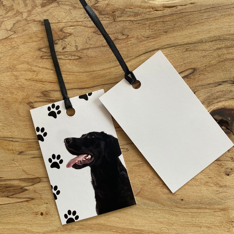 May include: A white gift tag with a black dog paw print design. The tag has a black ribbon attached and a hole punched in the top. The dog is a black Labrador Retriever with its tongue sticking out. The tag is blank for writing a message.