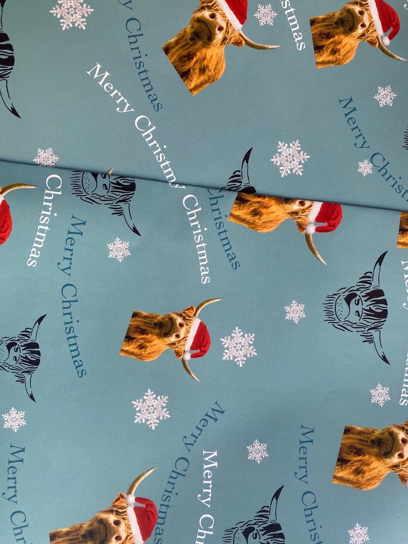 Op de afbeelding: Blauw inpakpapier met een herhalend patroon van Schotse Hooglanders met kerstmanmutsen en sneeuwvlokken. De tekst "Merry Christmas" wordt herhaald in het hele patroon.