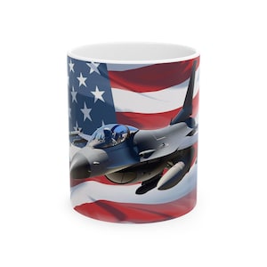 Pode incluir: Caneca de cerâmica branca com um caça cinzento contra um fundo da bandeira americana. O design inclui listras vermelhas e brancas e um campo azul com estrelas brancas. Uma caneca com tema patriótico.