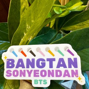 May include: A sticker with the text "BANGTAN SONYEONDAN BTS" in purple and white, featuring colorful microphone illustrations. The sticker is set against a backdrop of green leaves.