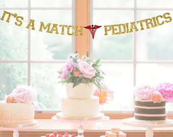Könnte beinhalten: Ein weißer Tisch mit einem goldenen Glitzerbanner, auf dem "It's a Match Pediatrics" mit einem roten medizinischen Symbol steht. Es gibt drei Kuchen auf Kuchentellern und einen Teller mit Macarons auf einem Kuchenteller. Der Tisch ist mit rosa Blumen dekoriert.