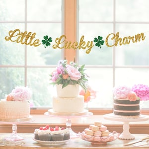 May include: A gold glitter banner with green shamrock accents reads "Little Lucky Charm".  The banner is hanging above a table with three cakes, cupcakes, and macarons. The cakes are on white cake stands with pink bases. The cupcakes are on a white plate with a pink base. The macarons are on a white plate with a pink base. The table is white with a wooden top.