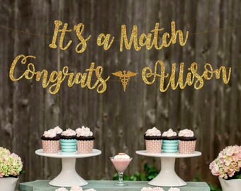 May include: Gold glitter banner with the text "It's a Match Congrats Allison" and a medical symbol. The banner is hanging above a table with cupcakes and other decorations.